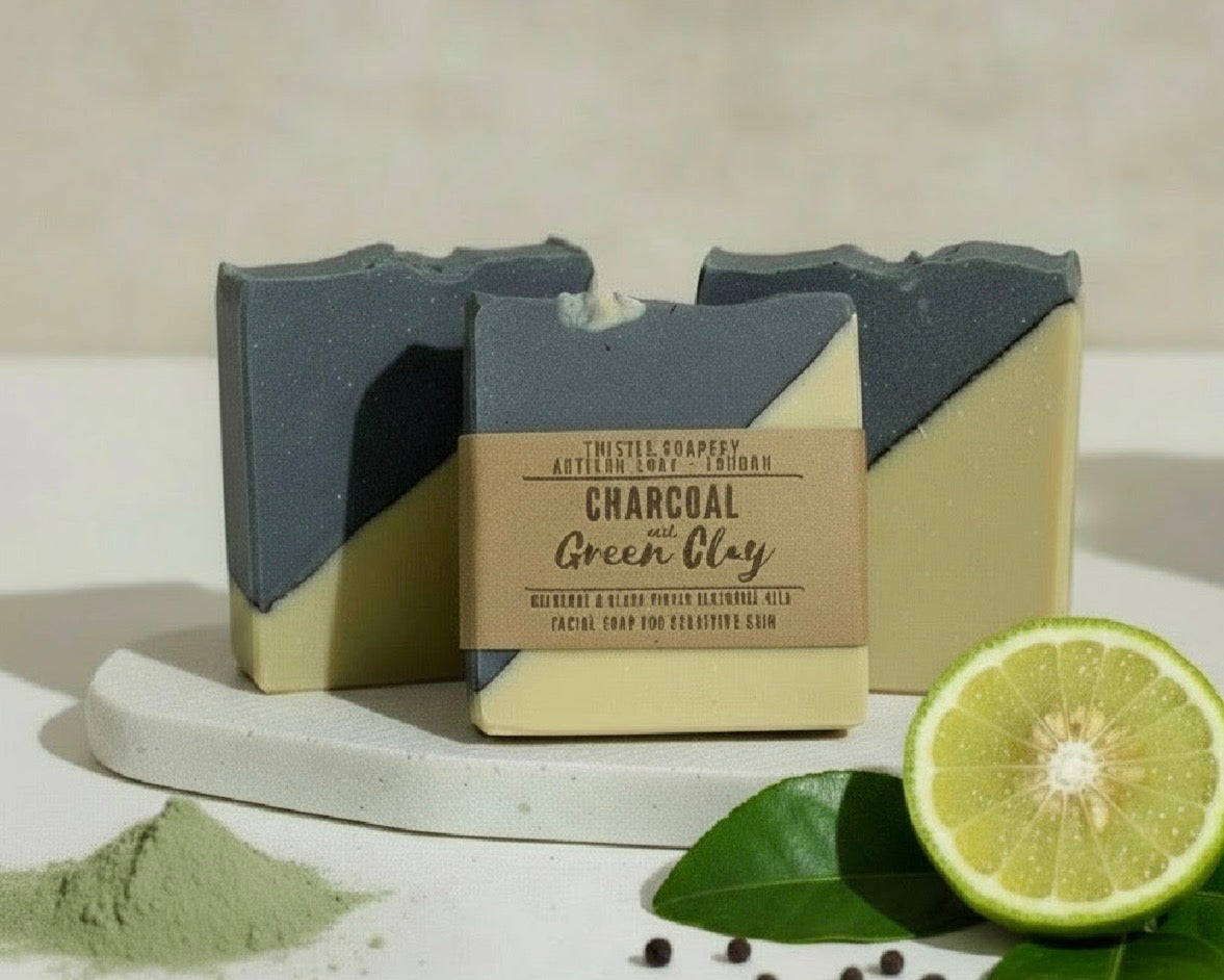Three bars of charcoal and green clay soap with a label, surrounded by lime and green clay on a light background.